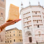 Holding a piece of Parmesan cheese on the Parma main square background in Italy. Parmesan is produced mostly in the province of Parma and was named after that producing region
